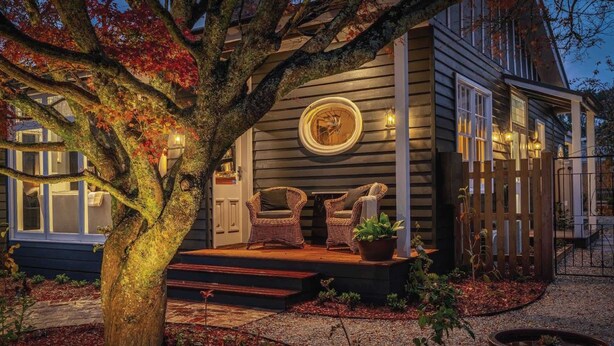 Una casa con porche, dos sillas de mimbre, un ventanal circular y un árbol con hojas otoñales.
