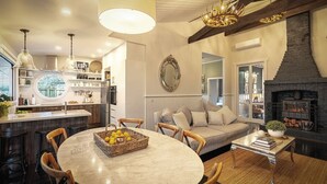 Una sala de estar acogedora con chimenea, una mesa de comedor redonda con sillas, un sofá con cojines y una zona de cocina con un ventanal redondo y estanterías.