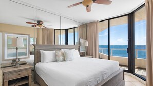 Une chambre d’hôtel dotée d’un grand lit, d’un ventilateur de plafond, d’une table de chevet avec une lampe et offrant une vue sur l’océan grâce à de grandes fenêtres.