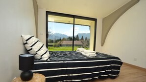 A bedroom with a large window, a bed with striped bedding, a bedside lamp, and a small wooden stool. - Gîte Trieves/ Gîte La Passerelle / Gîte Lavars (Lavars)