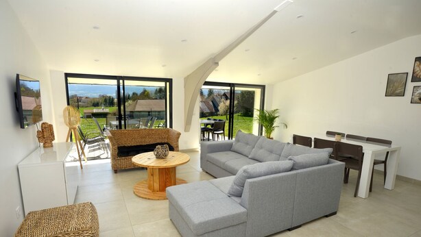 A modern living room with a grey sectional sofa, a round wooden coffee table, wicker furniture, and a dining area with a white table and brown chairs. - Gîte Trieves/ Gîte La Passerelle / Gîte Lavars (Lavars)