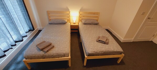 Two single beds with wooden frames and striped mattresses, a nightstand with a lamp, and a window with curtains.