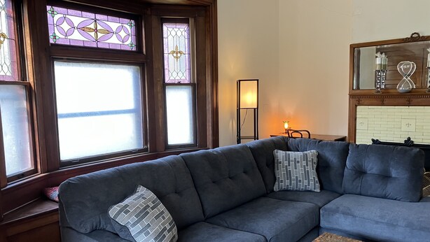 Ein Wohnzimmer mit einem dunkelgrauen Ecksofa, gemusterten Kissen, einem Teppich mit Muster und Buntglasfenstern.