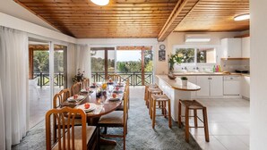 A dining area with a wooden table and chairs, a kitchen with white cabinets, and large windows offering a view of greenery. - Platres Forest Panorama (Platres)