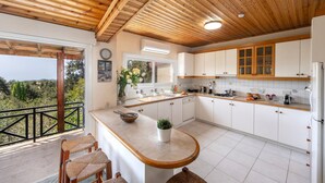 A kitchen with wooden ceiling beams, white cabinets, a large window, and a balcony with wicker chairs. - Platres Forest Panorama (Platres)