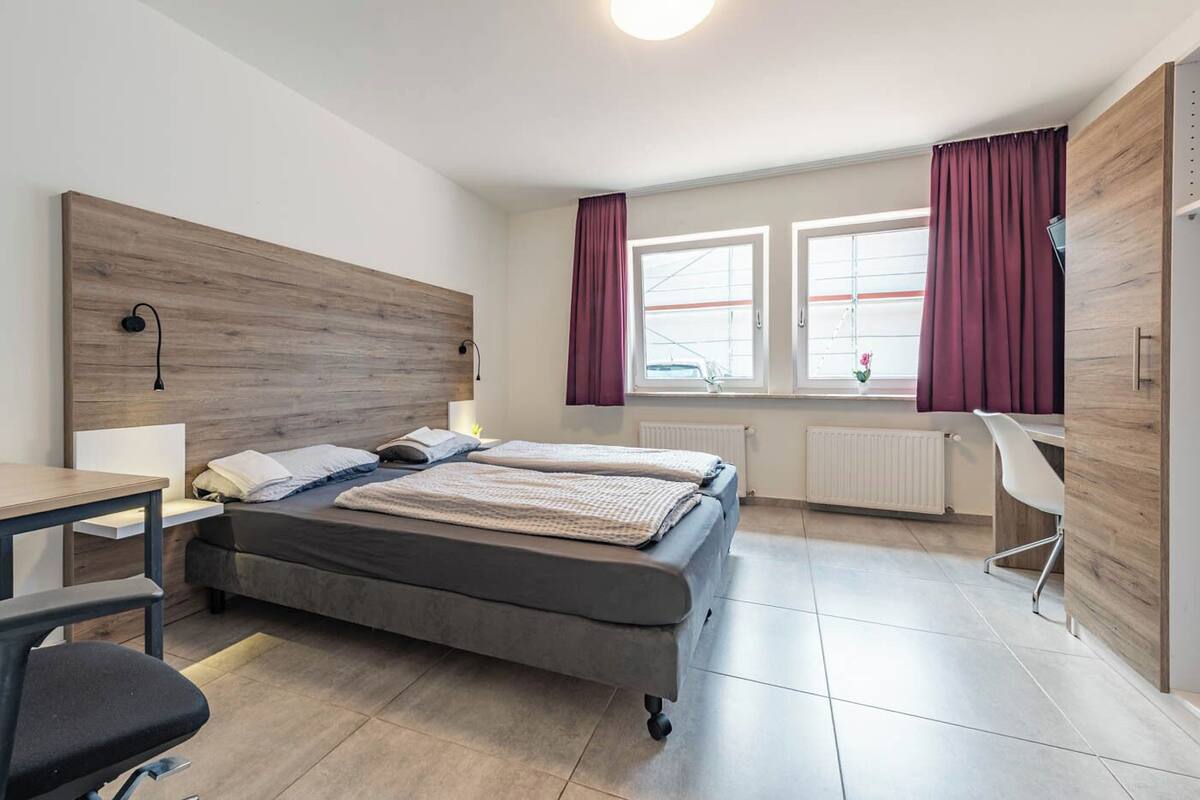 A modern hotel room with a bed, a desk, and a chair. There are two windows with curtains, a radiator, and a wooden headboard.