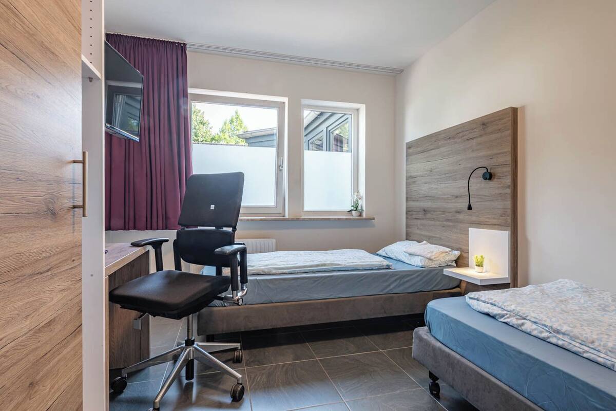 A compact hotel room with a desk, two single beds, a chair, and a window with curtains.