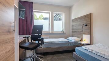 A compact hotel room with a desk, two single beds, a chair, and a window with curtains.