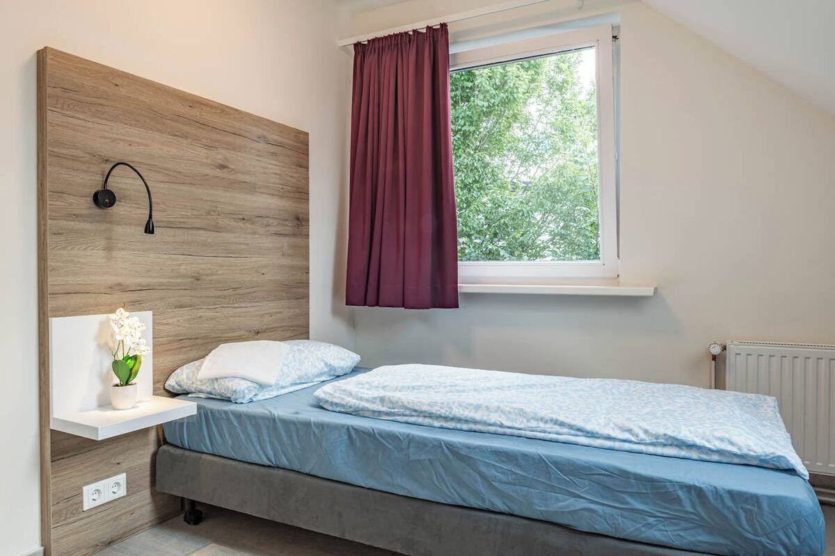 A bedroom with a bed, a nightstand, a wall-mounted lamp, a window with curtains, and a radiator.