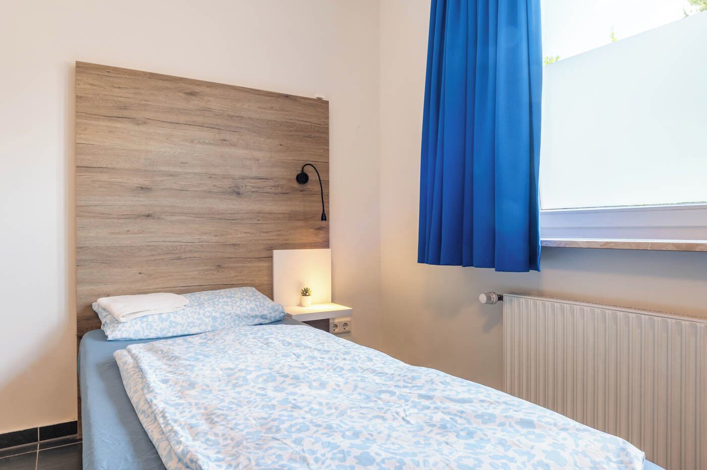 Ein Schlafzimmer mit einem hölzernen Kopfteil, einem Nachttisch mit Lampe, einem Fenster mit blauen Vorhängen und einer Heizung.