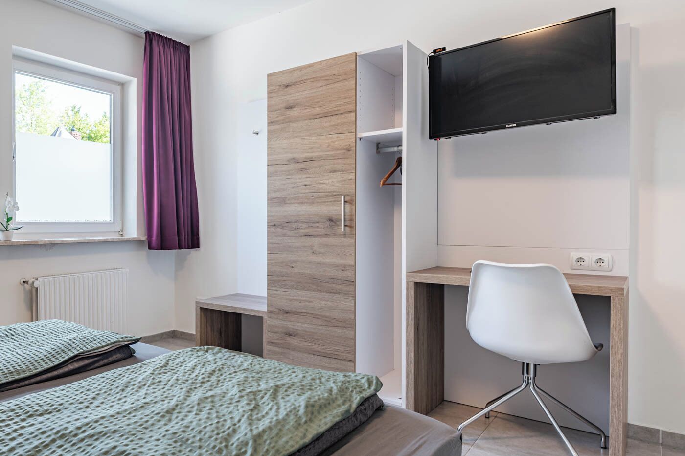 A modern bedroom with a bed, a desk, a chair, a TV, and a window with curtains.