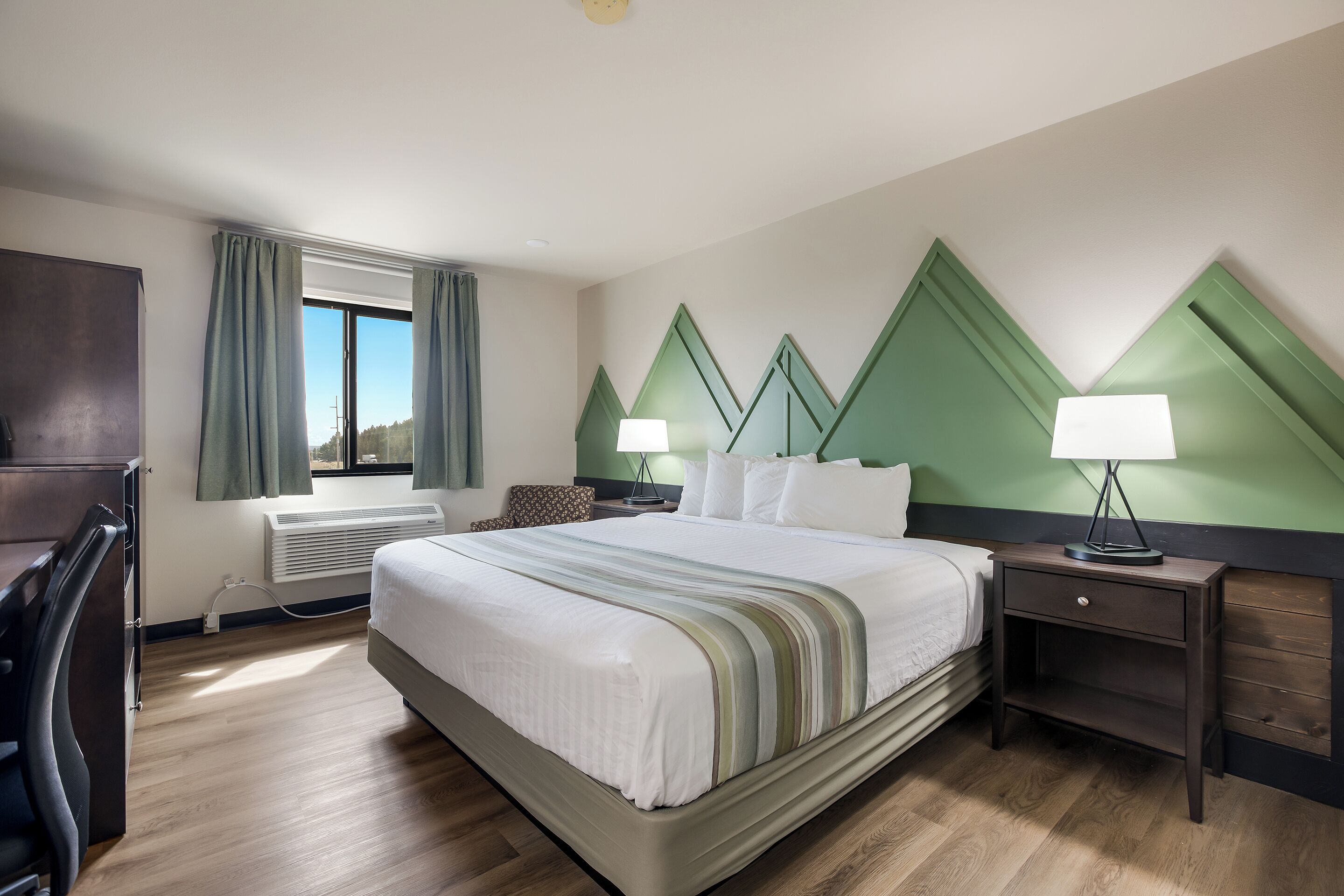 Habitación de hotel con una cama grande, dos mesitas de noche con lámparas, un escritorio, una silla y una ventana con cortinas.