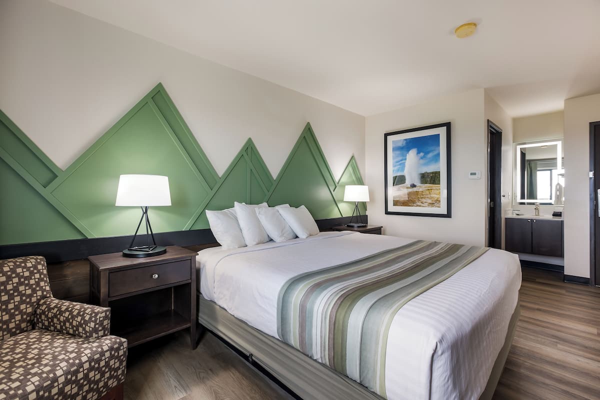 Een hotelkamer met een groot bed, twee nachtkastjes met lampen, een stoel, een schilderij aan de muur en een badkamer door een open deur zichtbaar.