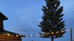 An outdoor deck with wooden flooring, metal chairs, and tables, surrounded by string lights and a large tree.