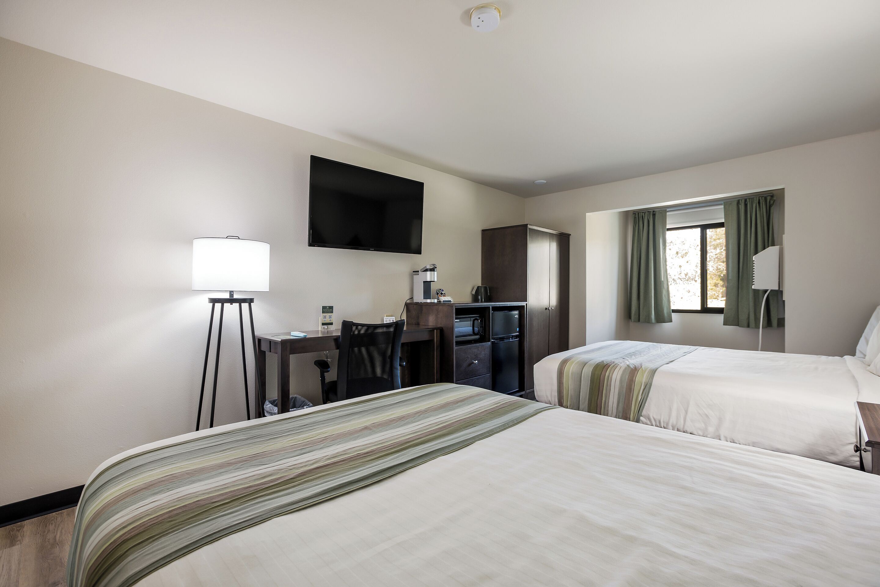 Ein Hotelzimmer mit zwei Betten, einem Schreibtisch mit Stuhl, einem Flachbildfernseher, einer Küchenzeile und einem Fenster mit Vorhängen.