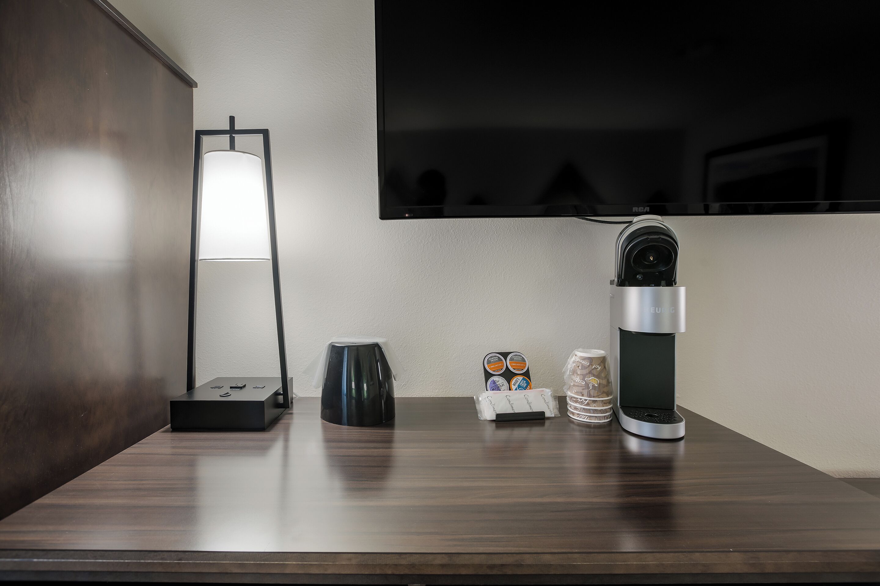 Una habitación de hotel moderna con un escritorio de madera oscura, una cafetera negra, dispensador de agua, portacápsulas de café y una cámara de seguridad en la pared.