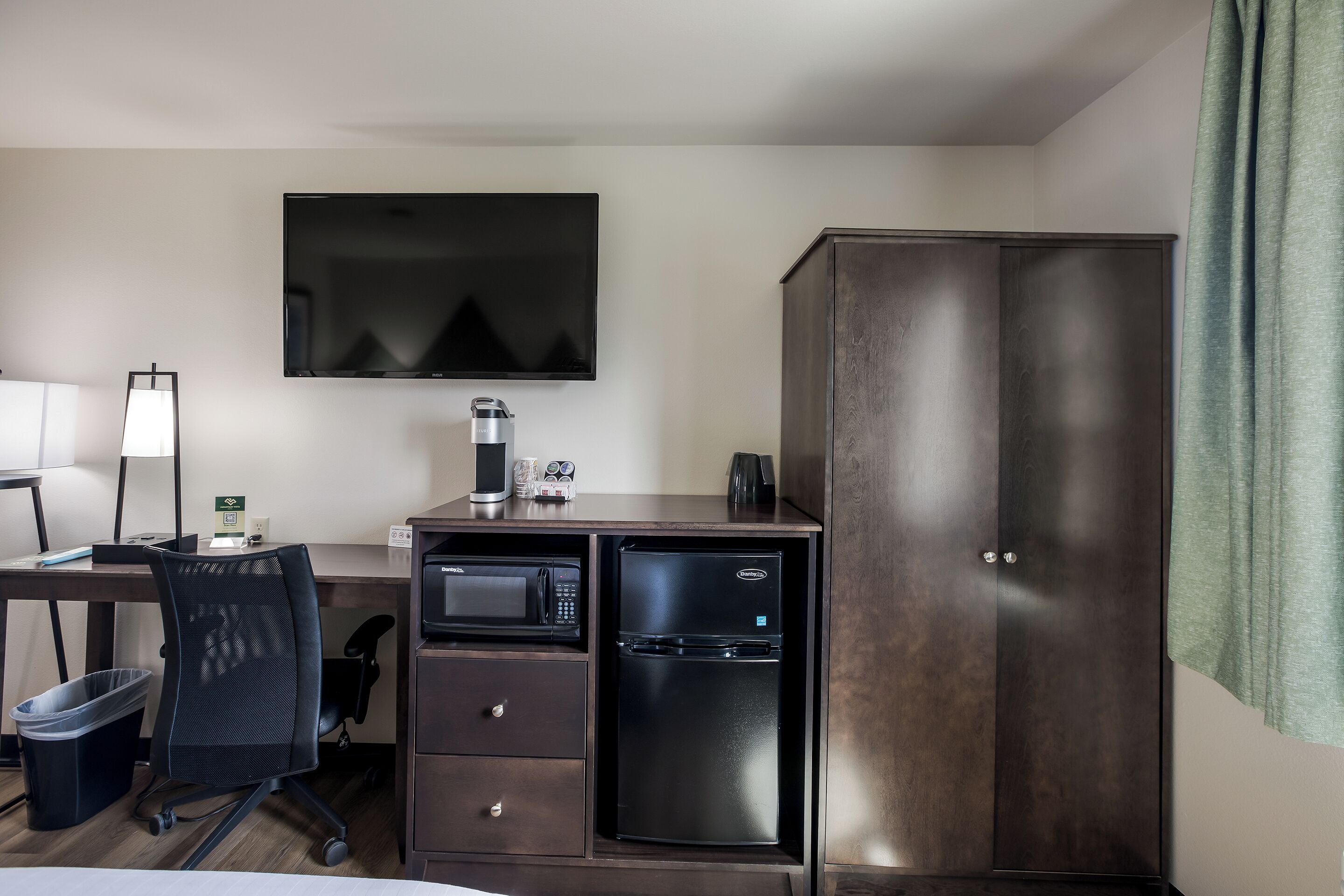 A hotel room with a desk, chair, TV, minibar, and wardrobe.