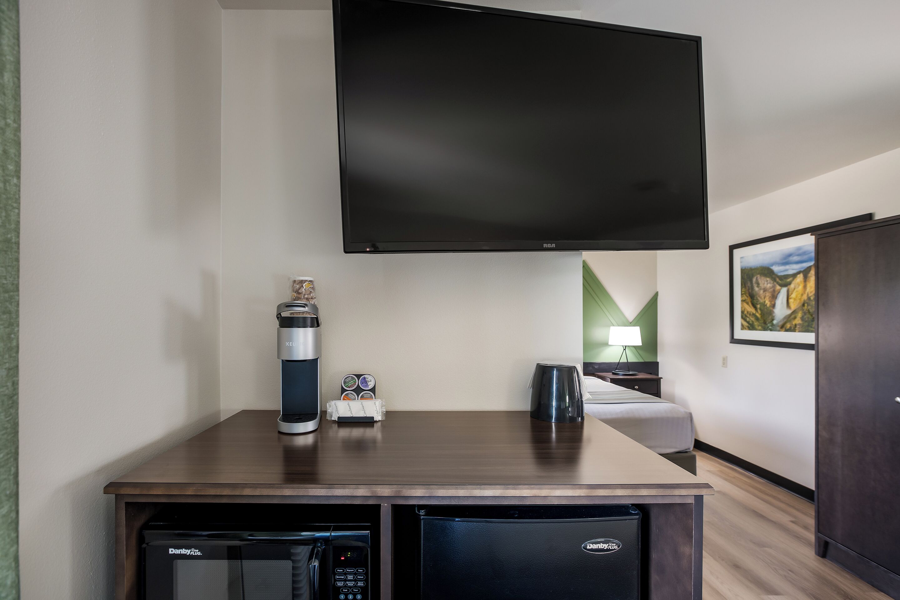 Habitación de hotel con televisor de pantalla plana en la pared, cafetera, refrigerador minibar y cama frente a una pared verde.