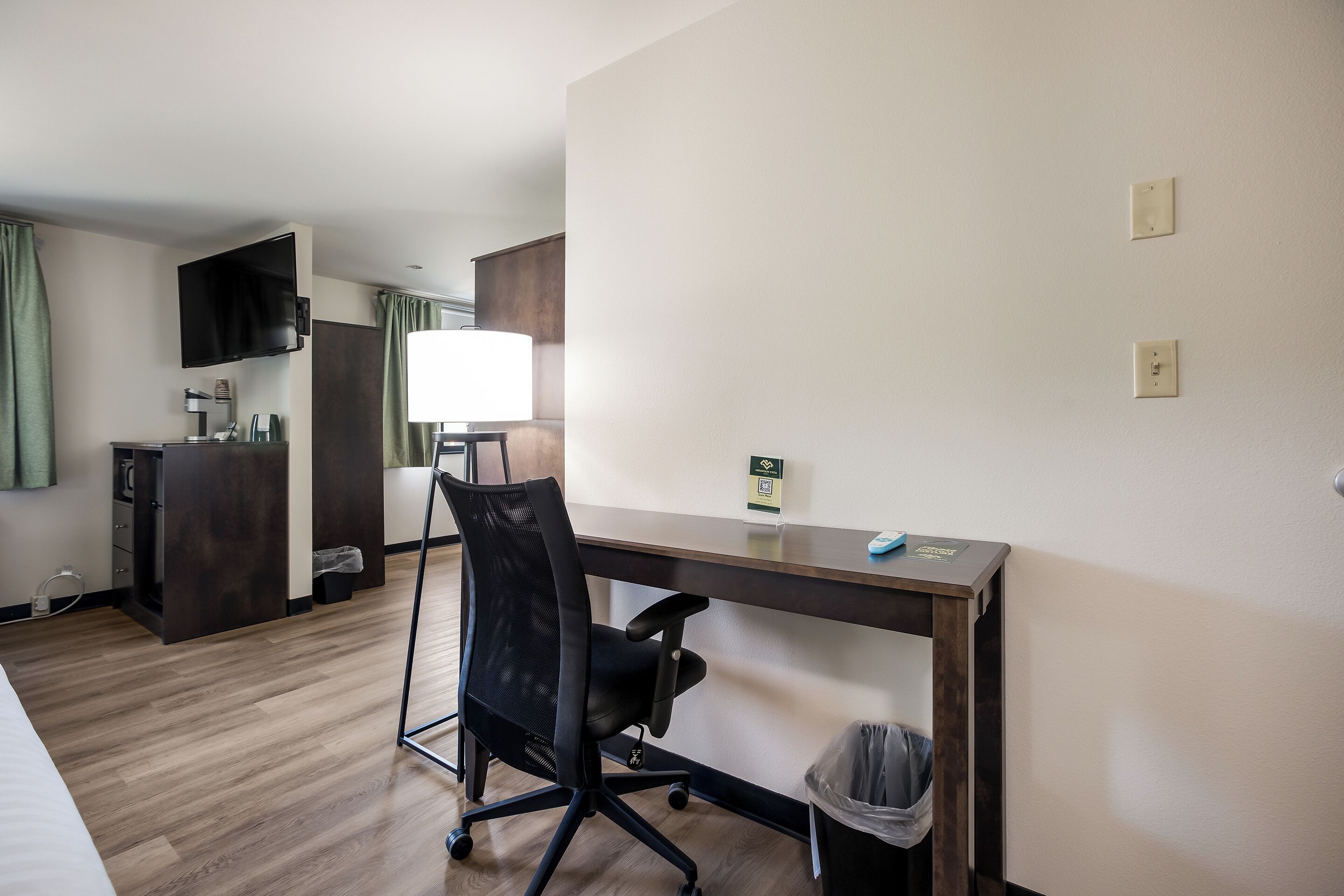 Habitación de hotel con piso de madera, un escritorio con silla, televisor montado en la pared y un cesto de basura.