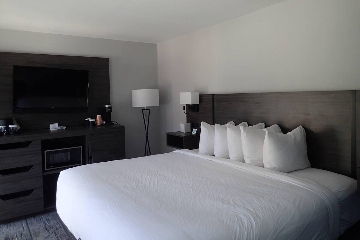 A hotel room with a large bed, a flat-screen TV mounted on the wall, a bedside table with a lamp, and a dresser with a mirror.