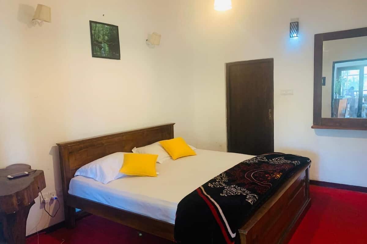 A single bed with a wooden frame, white bedding, and two yellow pillows. There is a dark-colored door to the right and a small table with a lamp to the left.
