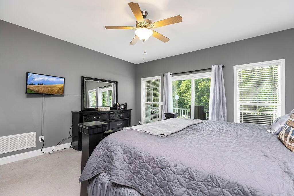 Une chambre avec un grand lit, un ventilateur de plafond, une télévision, une commode et une fenêtre avec des stores.