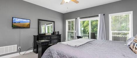 A bedroom with a large bed, a ceiling fan, a TV, a dresser, and a window with blinds.