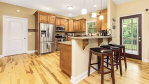 Una cocina con armarios de madera, electrodomésticos de acero inoxidable, una isla central con taburetes y vistas al exterior a través de una puerta.