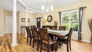 Comedor con mesa y sillas de madera, una araña, un reloj y una ventana con cortinas.