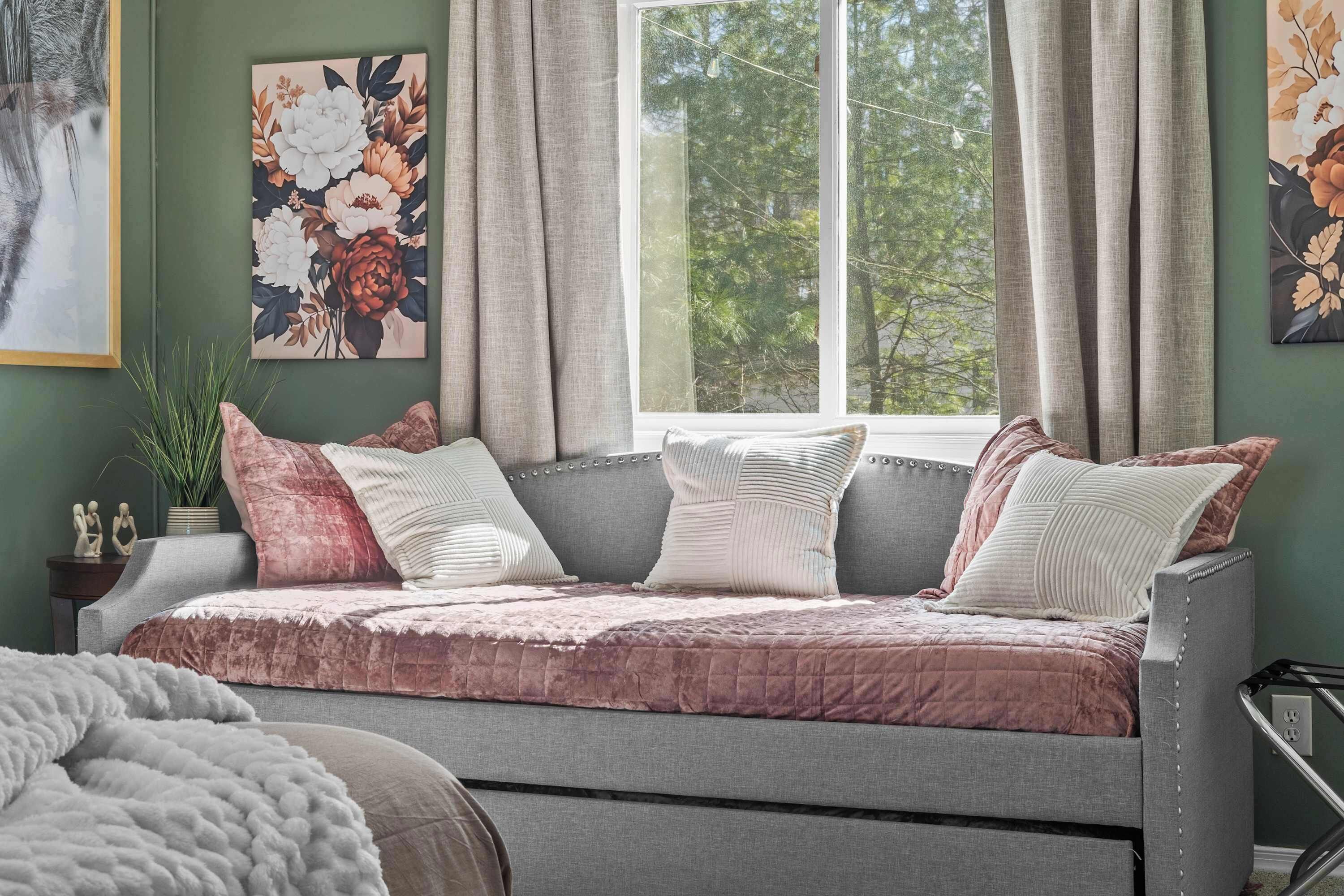 Un dormitorio acogedor con un sofá cama gris, obras de arte con motivos florales y vistas a árboles desde la ventana.