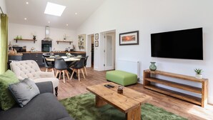 A modern living room with a sofa, coffee table, and TV. A kitchen with a dining area is visible in the background. - The Goose Shed (Canterbury)