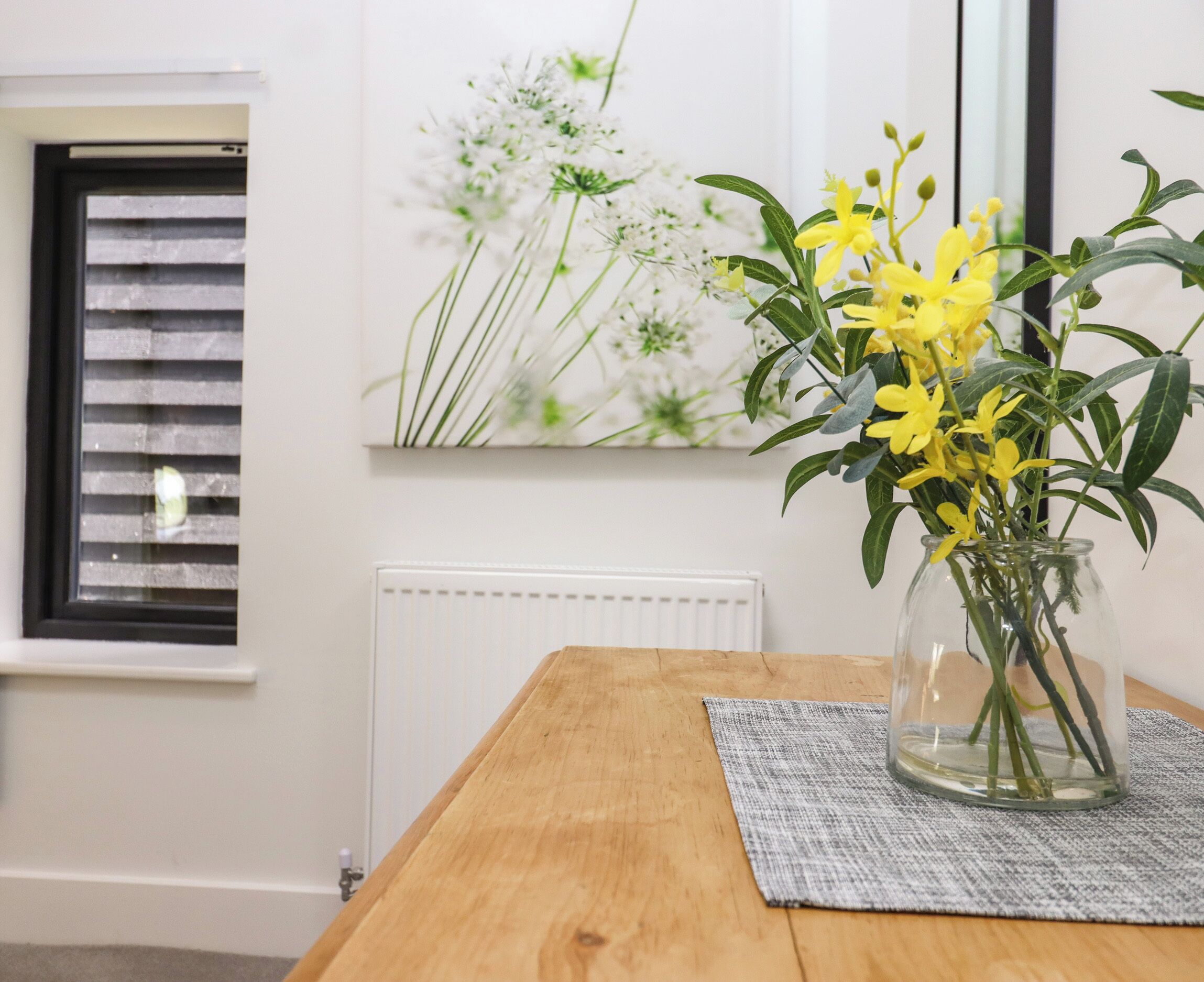 A wooden table with a glass vase containing yellow flowers, a gray placemat, a window with blinds, and a white radiator.