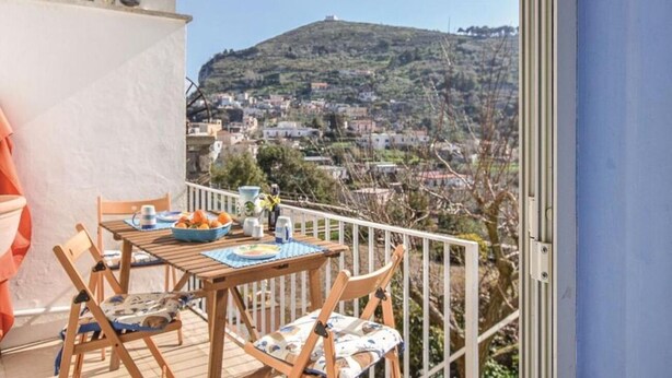 A balcony with a table set for two, overlooking a hillside town. - Delizia Caprese vacation home in Massa Lubrense with garden and Capri view (Massa Lubrense)