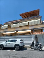 Un edificio de varios pisos con techo de tejas, un coche blanco estacionado al frente y una moto a un lado.