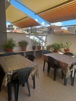 Un patio al aire libre con mesas y sillas, rodeado de plantas en macetas, bajo una zona sombreada y con vista a edificios al fondo.