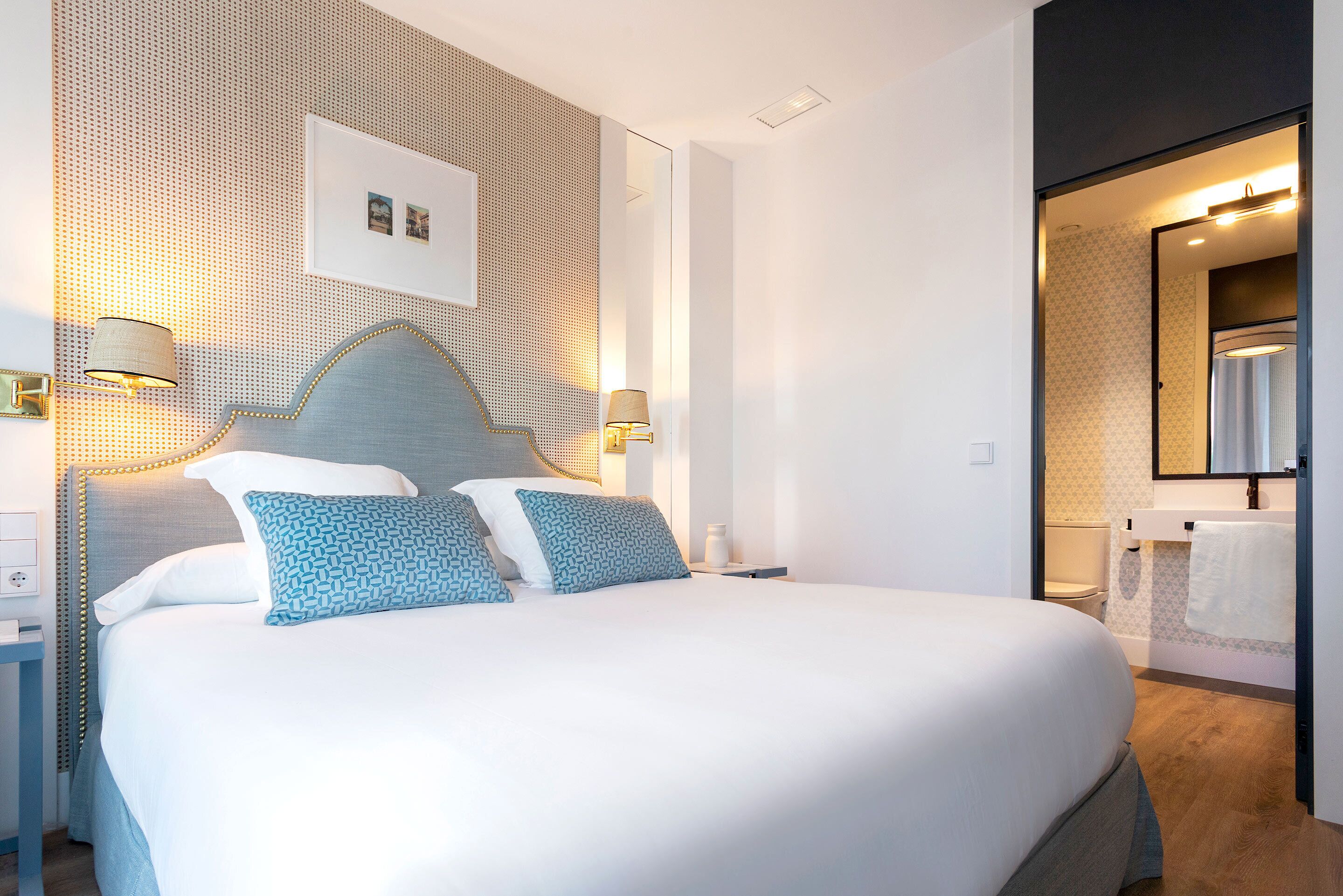 Una habitación de hotel moderna con una cama grande, dos lámparas de noche, un cabecero con diseño geométrico y un baño visible a través de una puerta abierta.