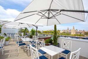 An outdoor seating area with white tables and chairs, large white umbrellas, and a view of a historic building across a river.