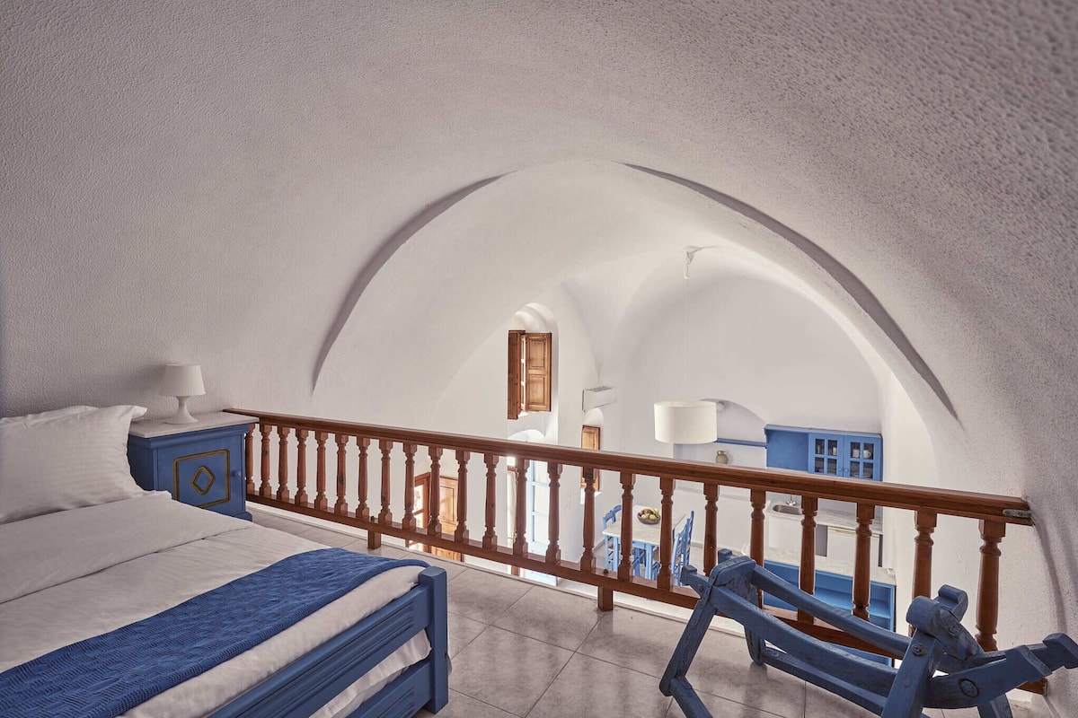 A bedroom with a bed, a nightstand, a wooden railing, and a view of a dining area.