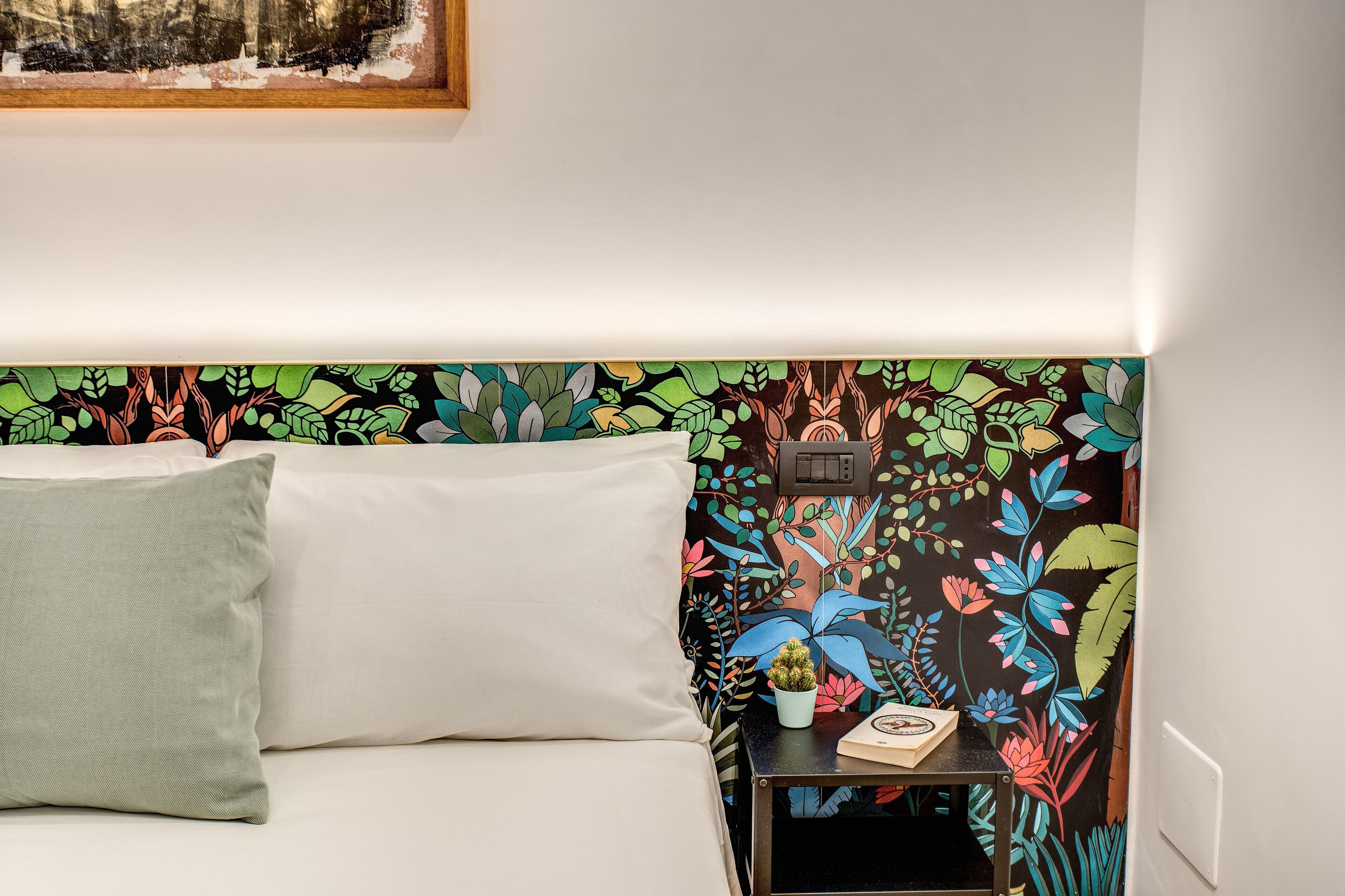 A neatly made bed with a green pillow, a small bedside table with a clock and a plant, and a colorful floral wallpaper.
