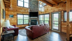 A log cabin interior with a stone fireplace, wooden walls, a leather sofa, and large windows.