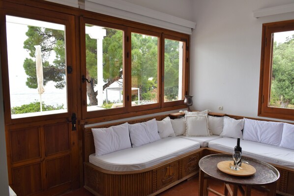 A room with a wooden bench sofa, a round table with a wine bottle, and large windows offering a view of trees and a body of water. - Villa Anthi (Skiathos)
