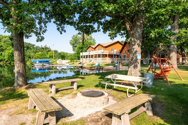 Enjoy a serene lakeside setting with a wooden deck and picnic tables, surrounded by mature trees and a two-story building with a balcony overlooking the water.