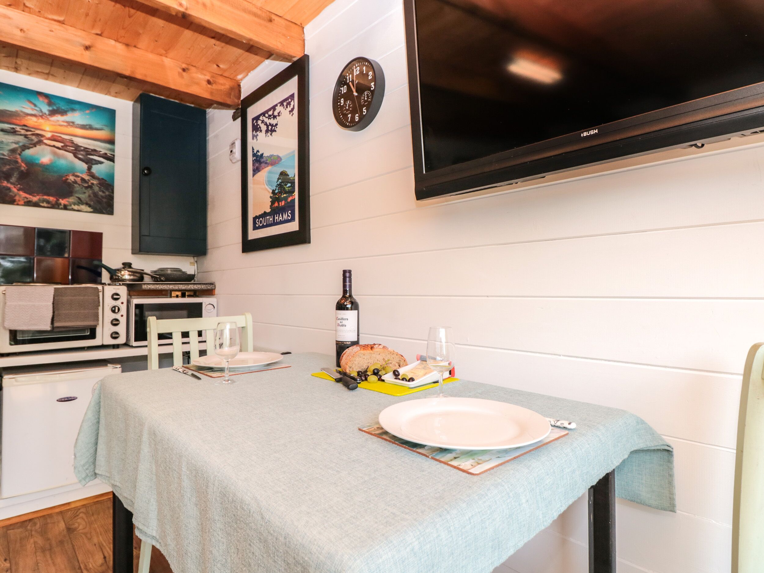 A kitchen with a table set for two, a clock, and a flat-screen TV mounted on the wall.