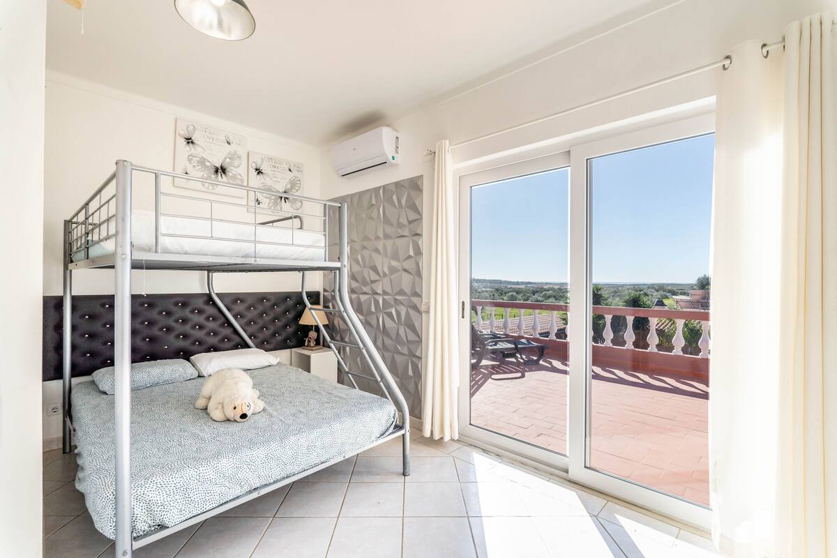 A bedroom with a bunk bed, a bedside table with a lamp, and a view of a balcony with outdoor furniture.