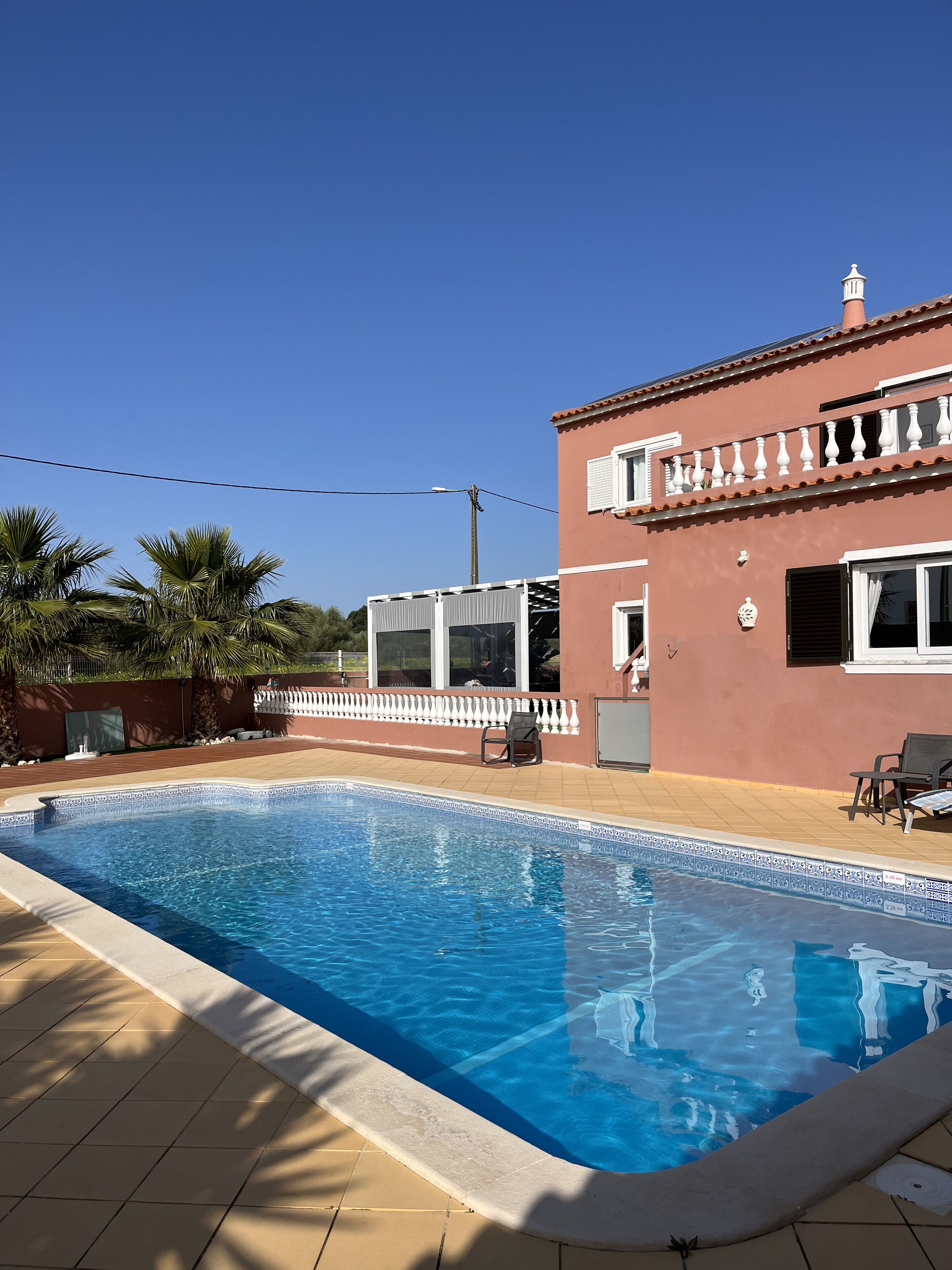Uma piscina com deck revestido, um edifício com varanda e palmeiras ao fundo.