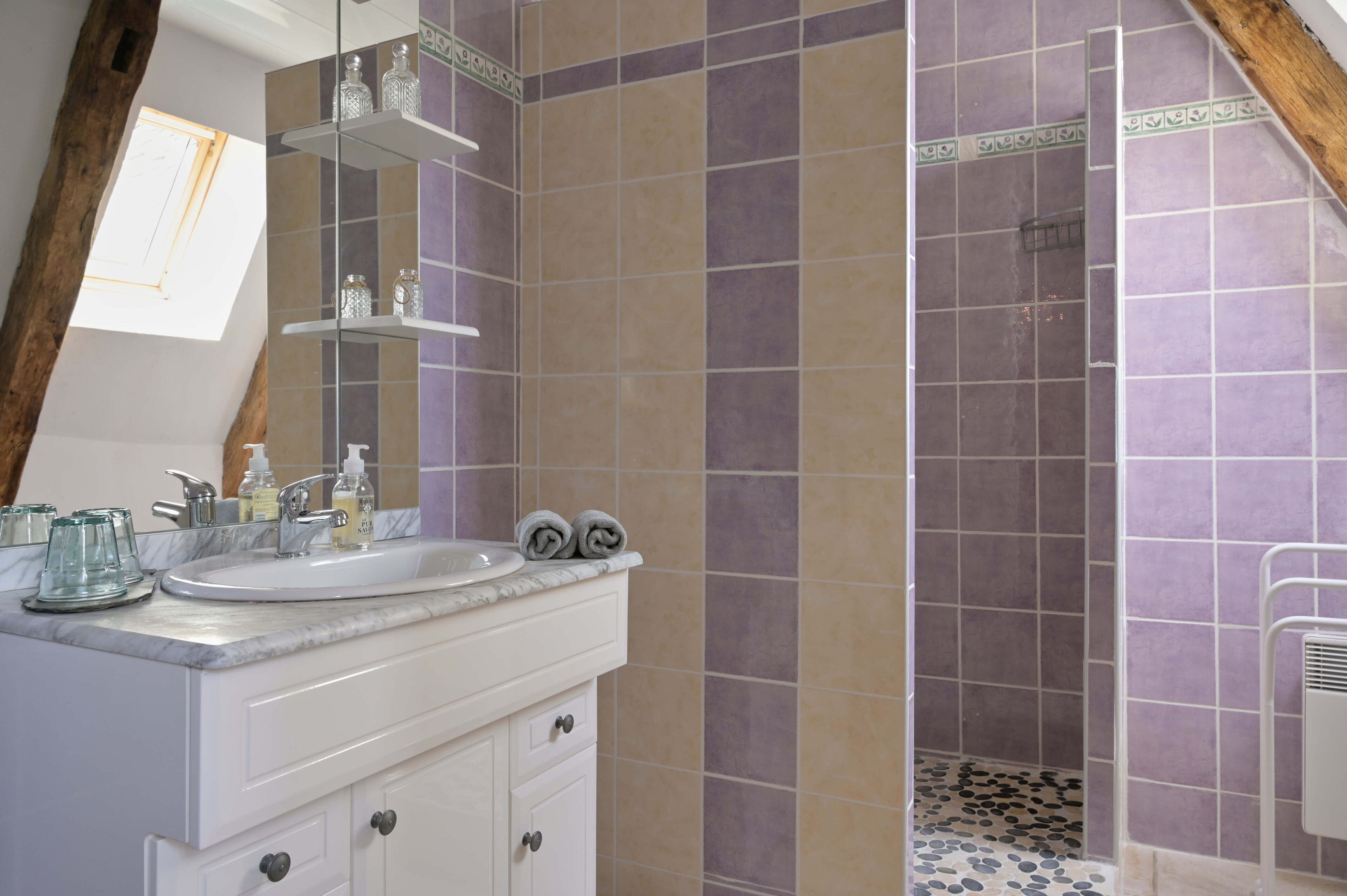 Une salle de bain avec un meuble-lavabo blanc, un évier et un miroir. Il y a des étagères au-dessus de l’évier et un espace douche avec des carreaux violets.