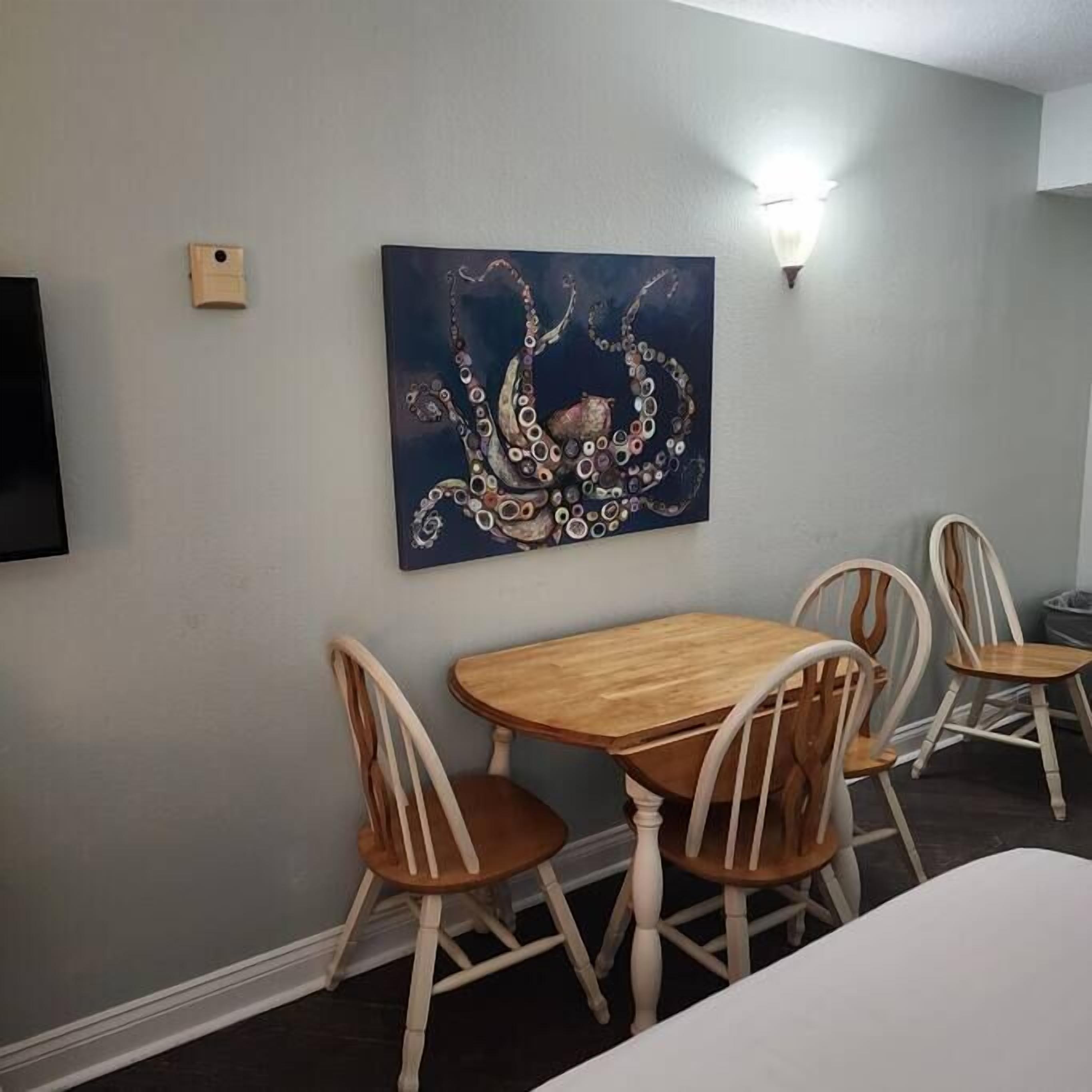 A dining area with a wooden table and four chairs, a wall-mounted painting, and a light fixture.