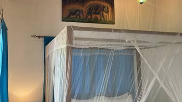 A room with a bed under a mosquito net, a ceiling fan, a painting of elephants, and a small table with a lamp.