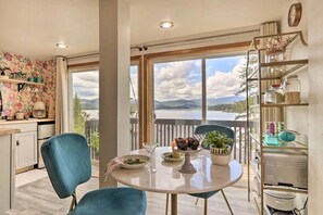 Un comedor con una mesa preparada para dos personas, vistas al exterior y una zona de cocina con papel tapiz floral y una estantería con frascos.