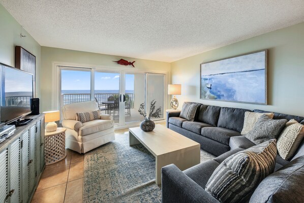 A living room with a sofa, armchairs, a coffee table, and a TV. There is a balcony with a view.